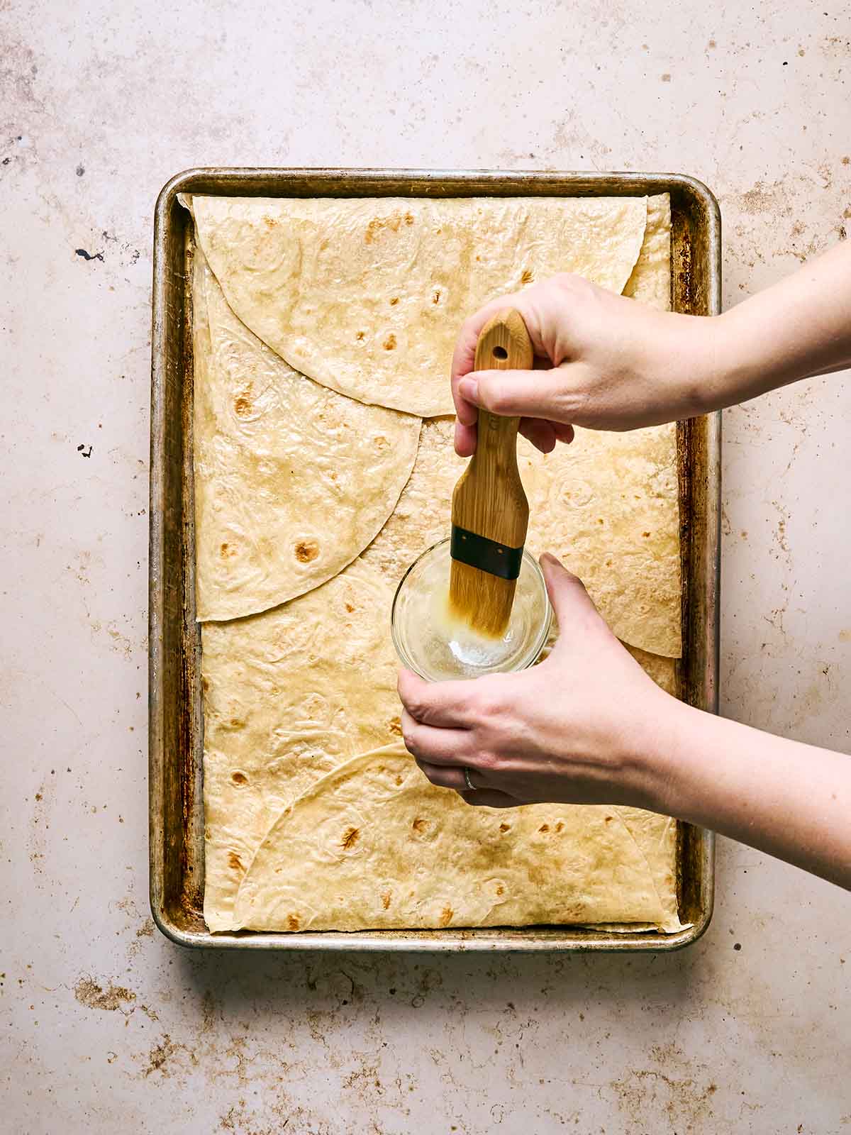 Two hands brush melted butter onto flour tortillas arranged in a single layer on a baking sheet. The scene is set on a light, textured surface.