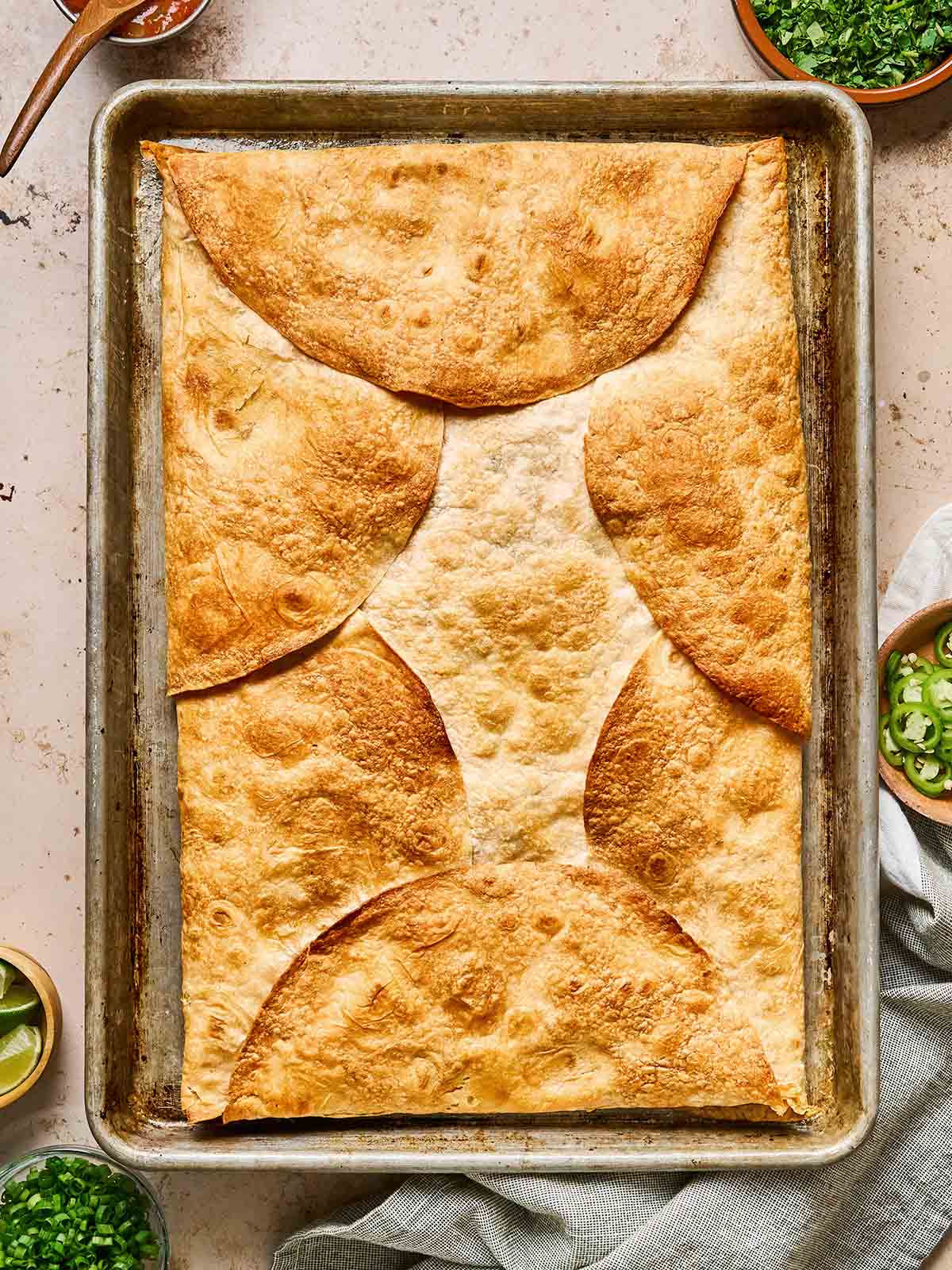 A rectangular baking sheet holds a golden, crispy baked dish topped with overlapping tortilla halves. Bowls with fresh chopped herbs and lime wedges are nearby on a light countertop.