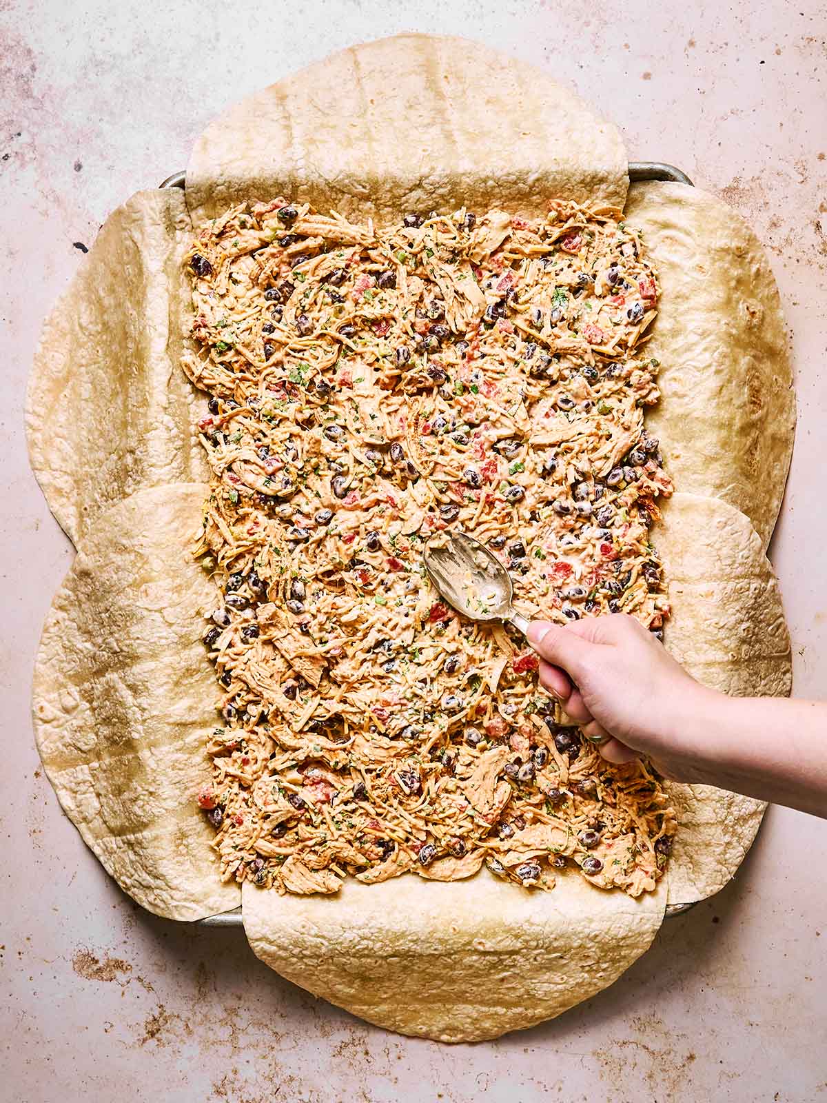 A hand spreads a mixture of shredded chicken, beans, cheese, and vegetables onto large flour tortillas arranged overlapping on a flat surface.