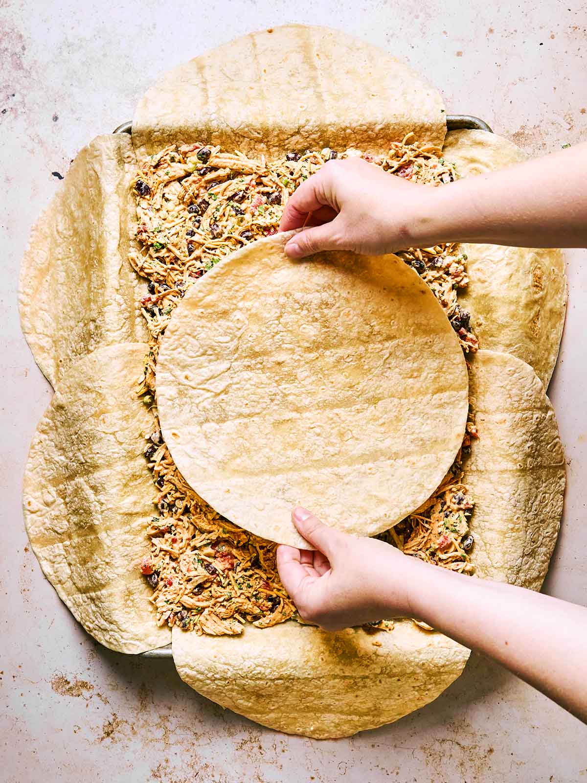 Two hands place a round tortilla on top of a layer of shredded chicken, black beans, and vegetables, surrounded by overlapping tortillas in a baking dish, preparing a layered casserole.