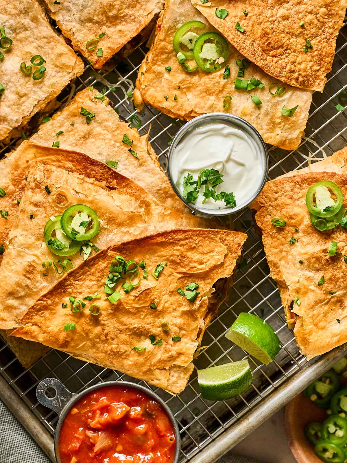 Golden brown quesadillas cut into triangles are arranged on a wire rack, topped with sliced jalapeños and green onions. Small bowls of sour cream and salsa, plus lime wedges, are placed beside them.