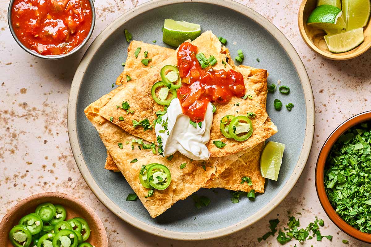A plate of quesadillas topped with sour cream, salsa, and sliced jalapeños, garnished with chopped cilantro. Lime wedges, salsa, jalapeños, and cilantro are in small bowls around the plate.