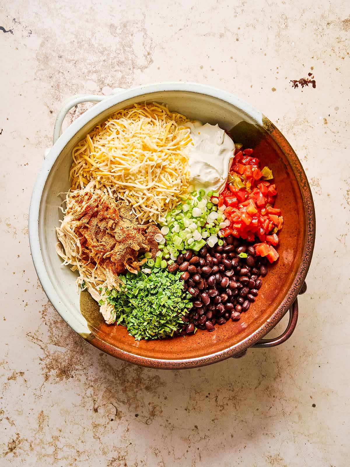 A large bowl on a light surface containing shredded cheese, sour cream, diced tomatoes, black beans, chopped green onions, cilantro, shredded chicken, and spices arranged in separate sections.