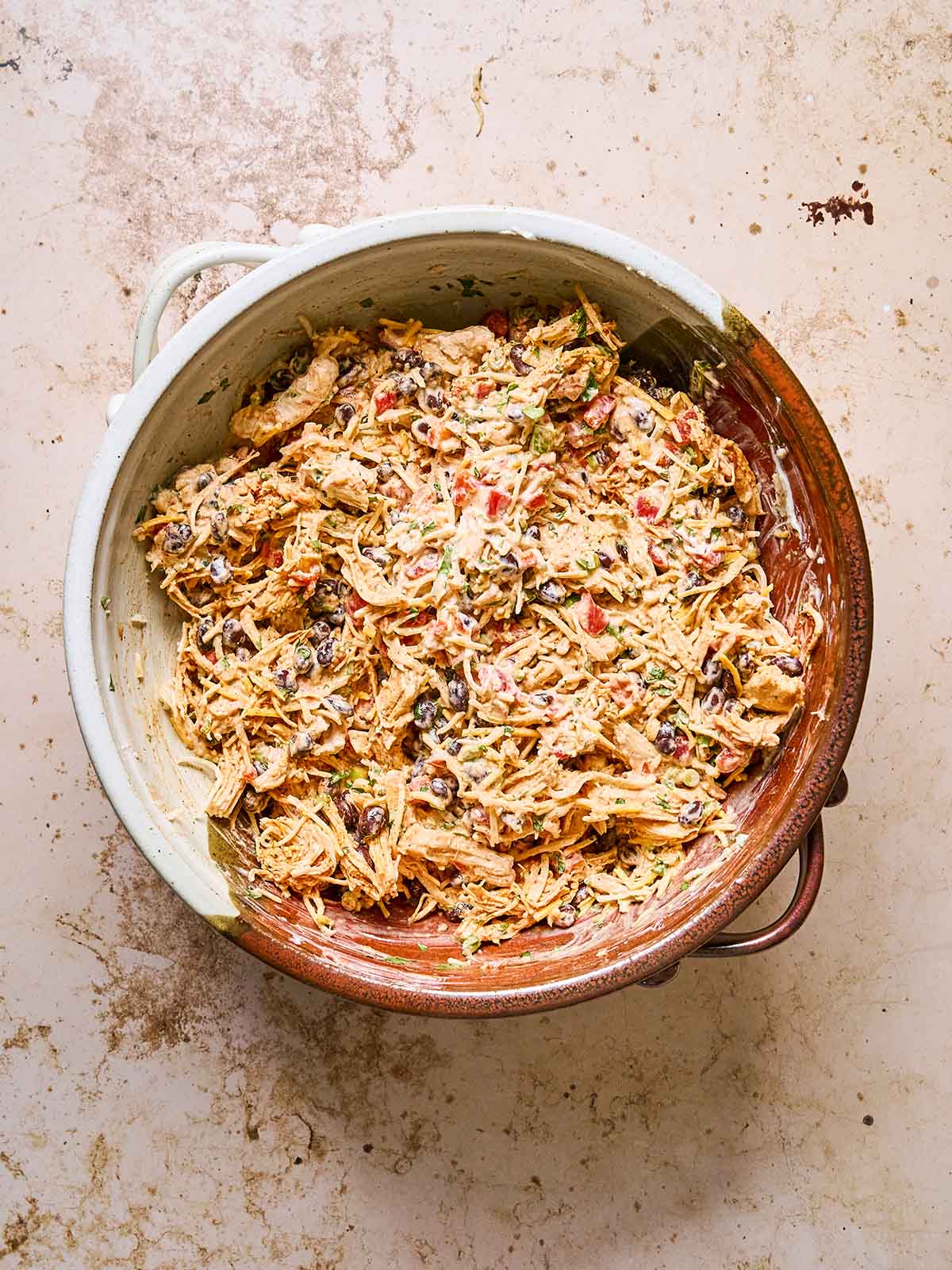 A large ceramic bowl filled with shredded chicken mixed with black beans, corn, chopped tomatoes, green herbs, and a creamy sauce, sitting on a light, textured surface.