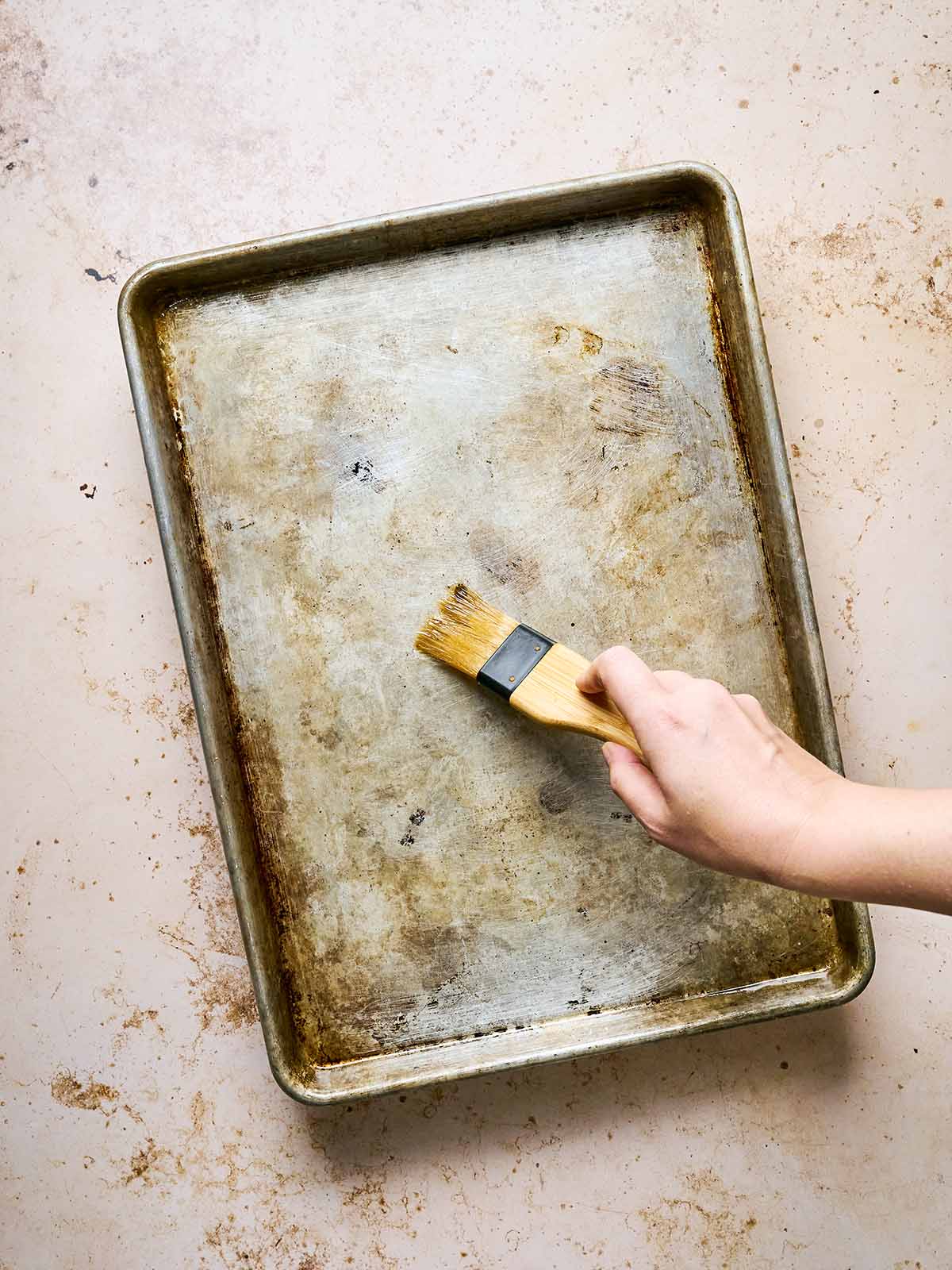 A hand holds a pastry brush, brushing the surface of a worn, metal baking sheet placed on a light, speckled countertop.