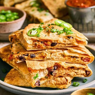 A stack of quesadilla wedges filled with beans, cheese, and vegetables, topped with sliced jalapeños and green onions, served on a plate with salsa and garnishes in the background.