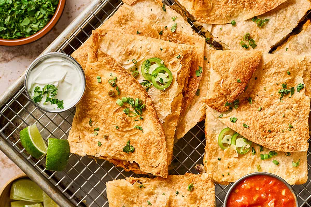 A tray of crispy quesadillas garnished with sliced jalapeños and chopped herbs, served with lime wedges, sour cream, and salsa in small bowls on the side.