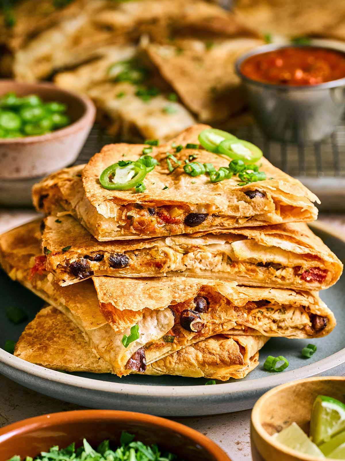 A stack of quesadilla wedges filled with beans, cheese, and vegetables, topped with sliced jalapeños and green onions, served on a plate with salsa and garnishes in the background.