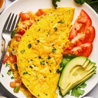 A cheesy omelette filled with diced vegetables is served on a white plate, garnished with chopped herbs. Slices of avocado, tomato, and fresh cilantro are on the side, with a fork and a checkered napkin nearby.