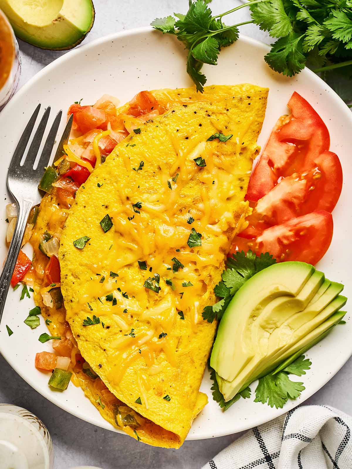 A cheesy omelette filled with diced vegetables is served on a white plate, garnished with chopped herbs. Slices of avocado, tomato, and fresh cilantro are on the side, with a fork and a checkered napkin nearby.