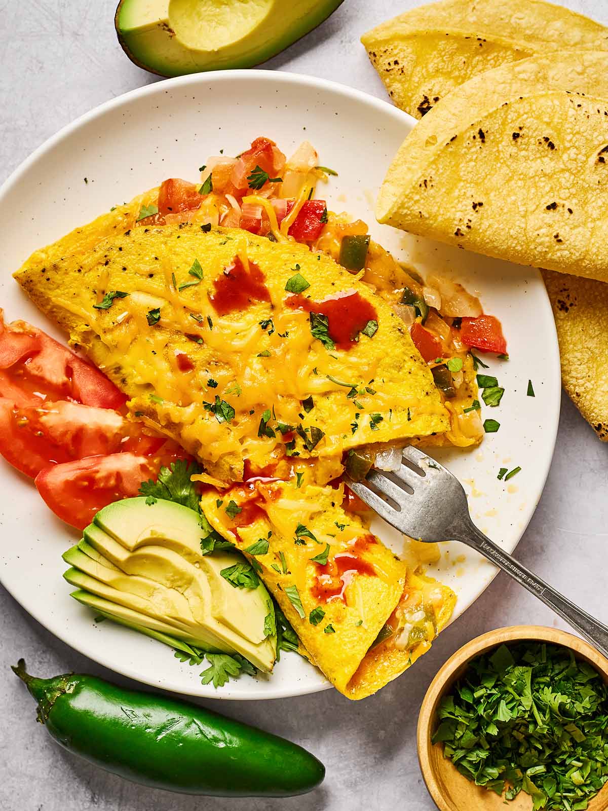 A plate with a cheesy omelette topped with chopped herbs, served with sliced avocado, tomato, and warm corn tortillas. A jalapeño, avocado half, and a bowl of chopped cilantro are nearby.