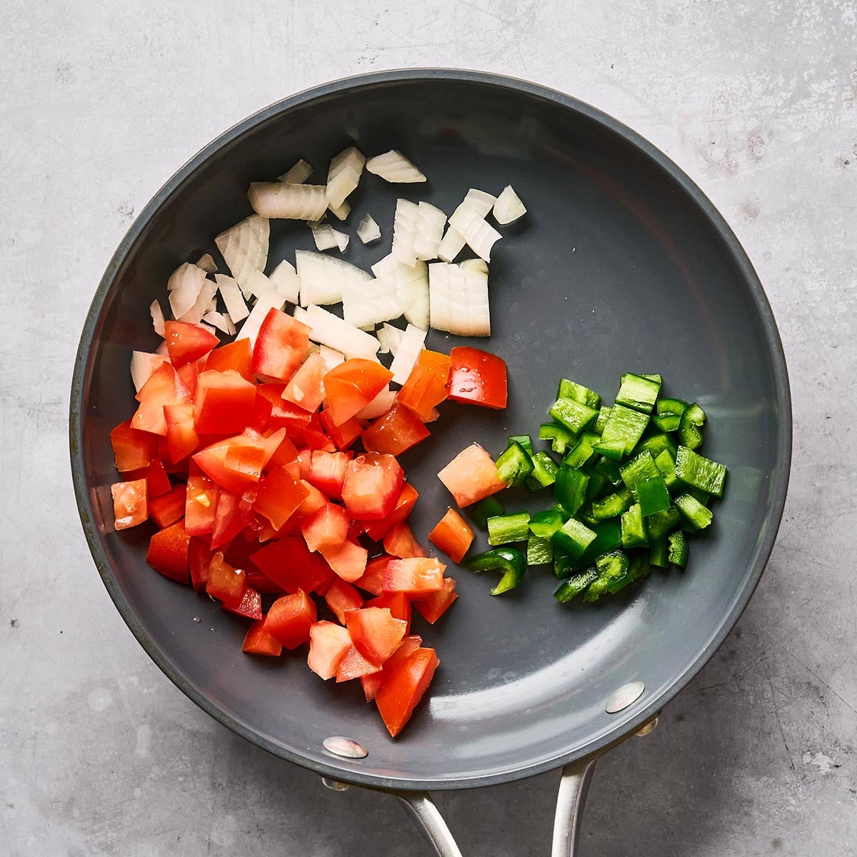 A frying pan contains chopped onions, tomatoes, and green bell peppers, each ingredient grouped separately on the pan’s surface, ready for cooking.