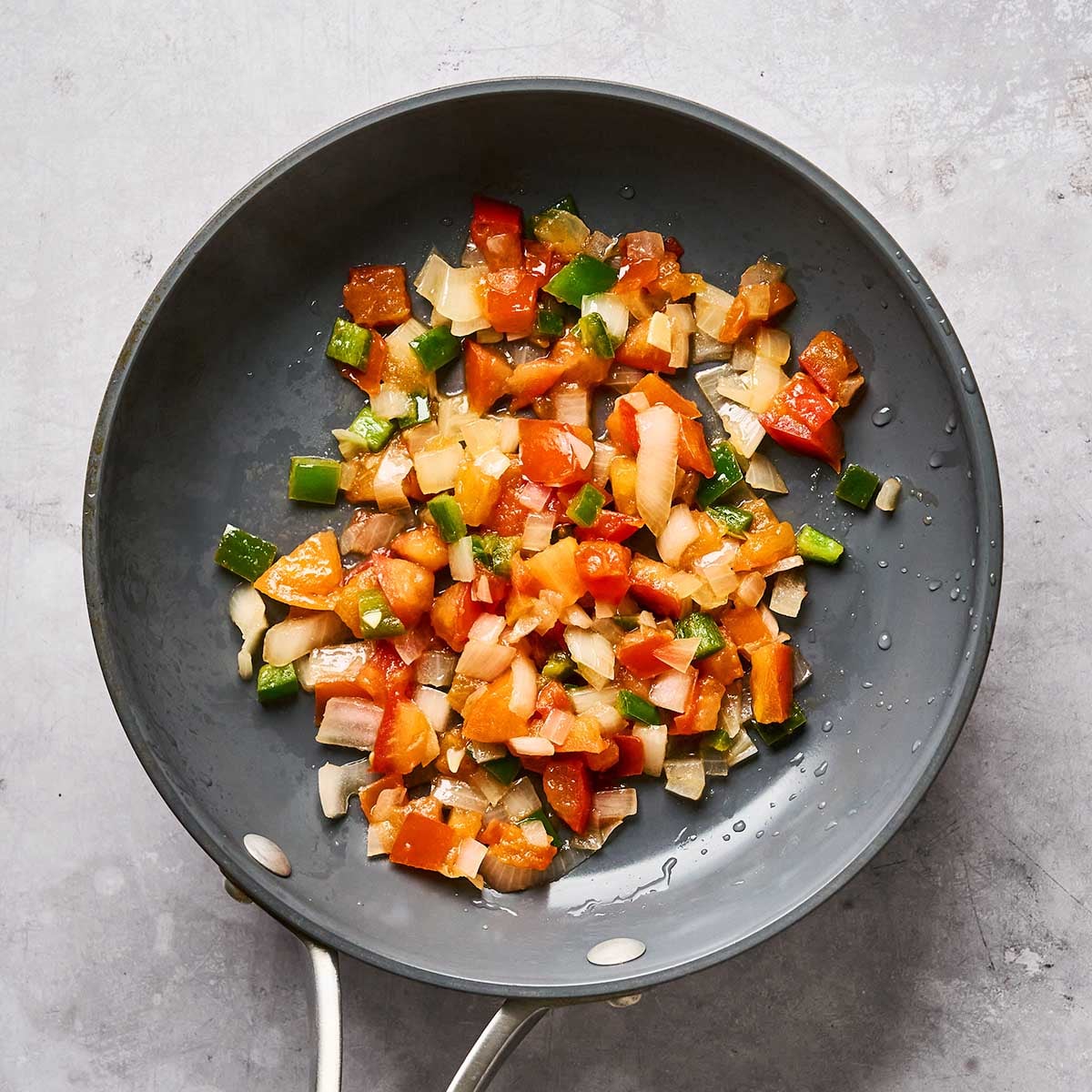A frying pan containing diced sautéed vegetables, including tomatoes, onions, and green peppers, sits on a light gray surface.