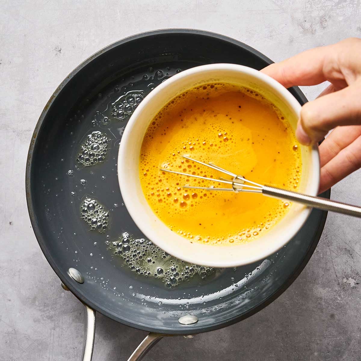 A hand pours a bowl of whisked eggs into a frying pan with melted butter on a gray countertop.
