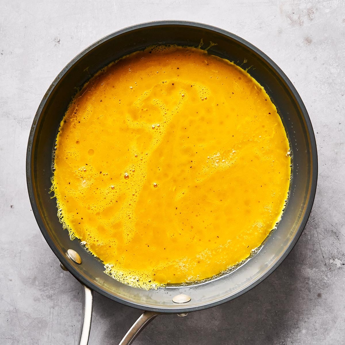 A nonstick frying pan with beaten eggs cooking in it, forming the base of an omelette, sits on a gray countertop. The eggs are partially set and appear bright yellow with a few bubbles.