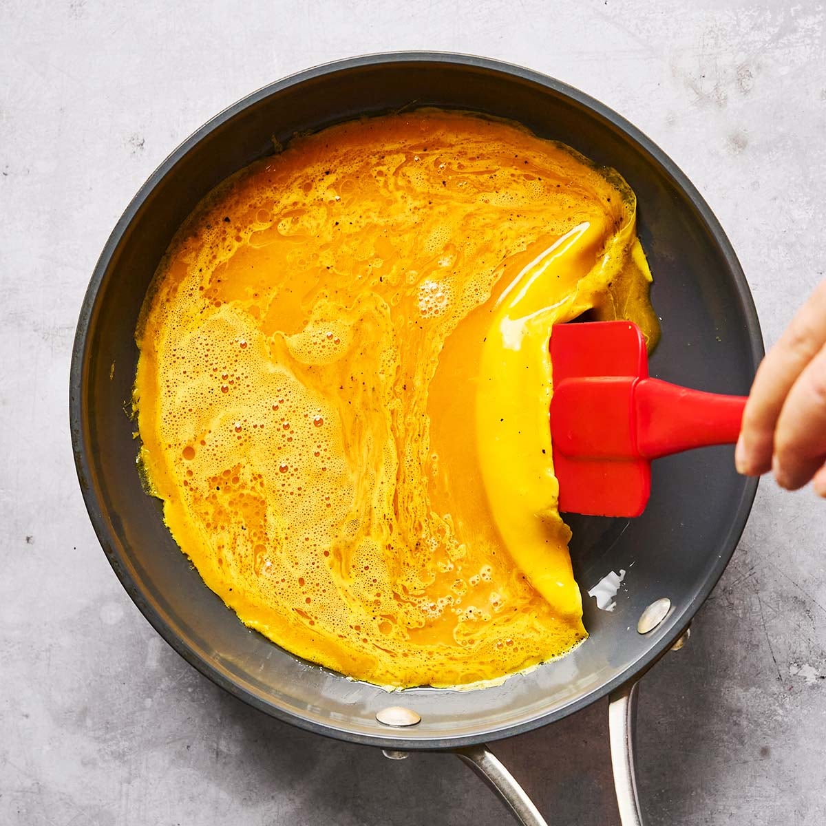 A hand uses a red spatula to fold a thin layer of yellow scrambled eggs or omelet in a nonstick frying pan on a gray surface.