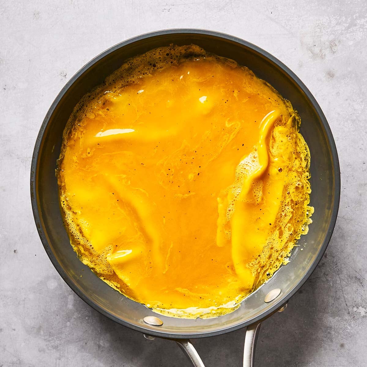 A black skillet on a gray surface contains partially cooked, bright yellow scrambled eggs, with some liquid areas still visible.
