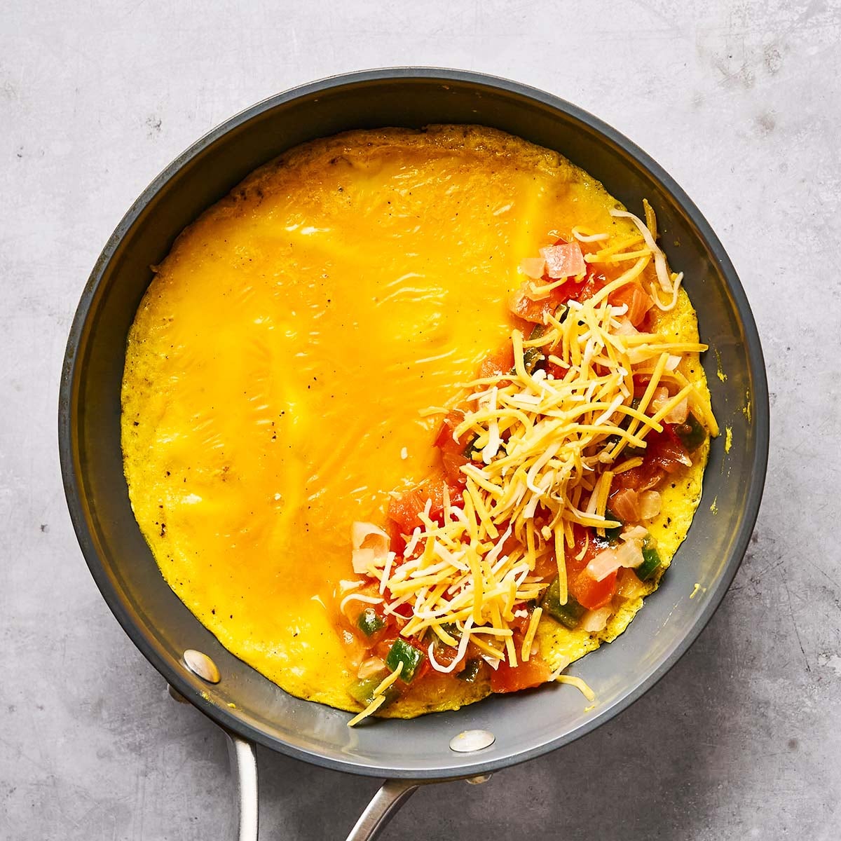 A frying pan with a cooked omelet, half covered in melted cheese, and the other half topped with shredded cheese, diced tomatoes, onions, and greens, on a gray surface.