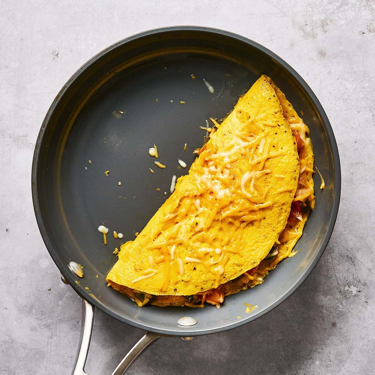 A cheese omelette folded in half sits in a nonstick frying pan, with melted cheese sprinkled on top and some cheese and fillings visible inside. The pan is placed on a light gray surface.