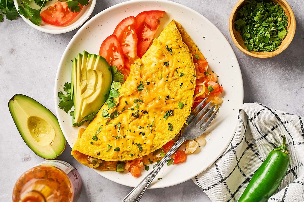A plate with a folded omelette garnished with chopped herbs, served with sliced avocado, tomato, cilantro, and a fork. Nearby are a bowl of chopped herbs, half an avocado, a napkin, hot sauce, and a green chili pepper.