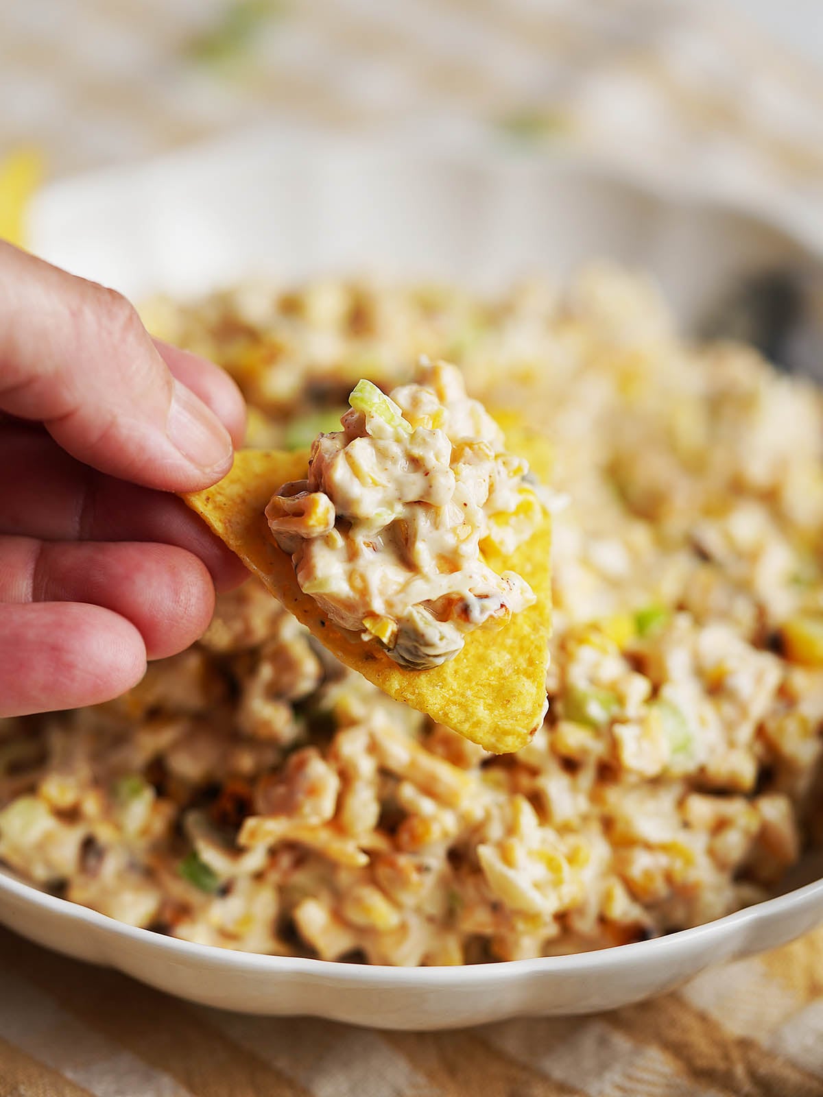 A hand holds a tortilla chip topped with creamy corn dip over a bowl filled with Street Corn Dip, featuring visible corn kernels and green bits.