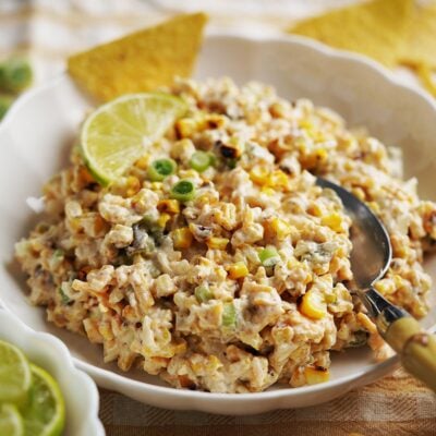 A bowl of creamy corn salad with chopped vegetables, garnished with lime slices and green onions, served with tortilla chips on the side. A spoon rests in the bowl.