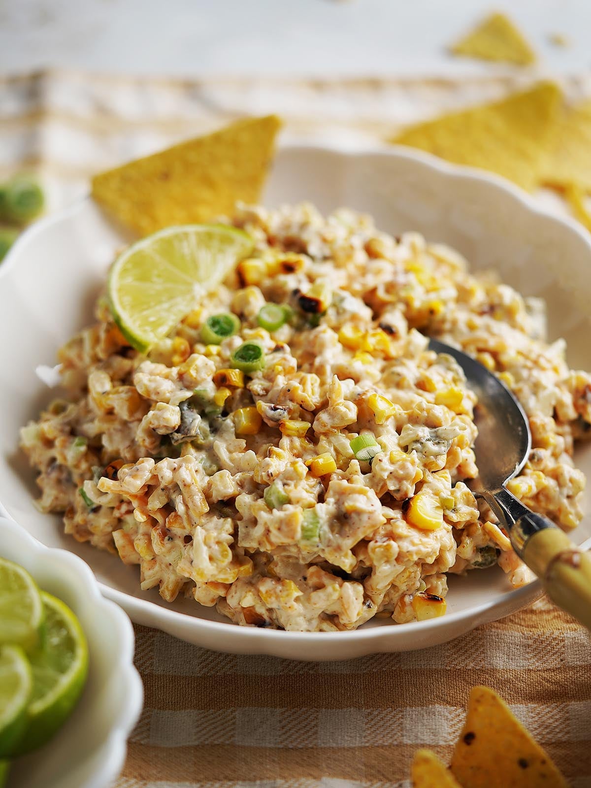 A bowl of creamy Street Corn Dip with chopped vegetables, garnished with lime slices and green onions, served with tortilla chips on the side. A spoon rests in the bowl.