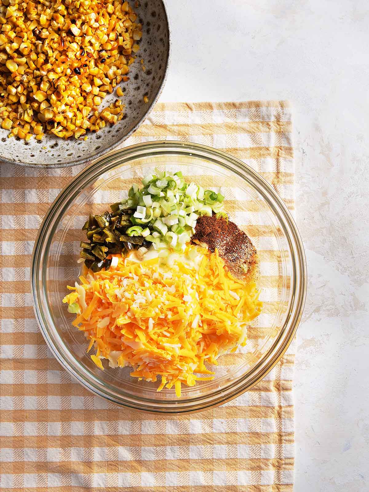 A glass bowl with shredded cheese, chopped green onions, diced jalapeños, mayonnaise, and seasonings sits on a checkered cloth. A dish of roasted corn is partially visible in the top left corner.