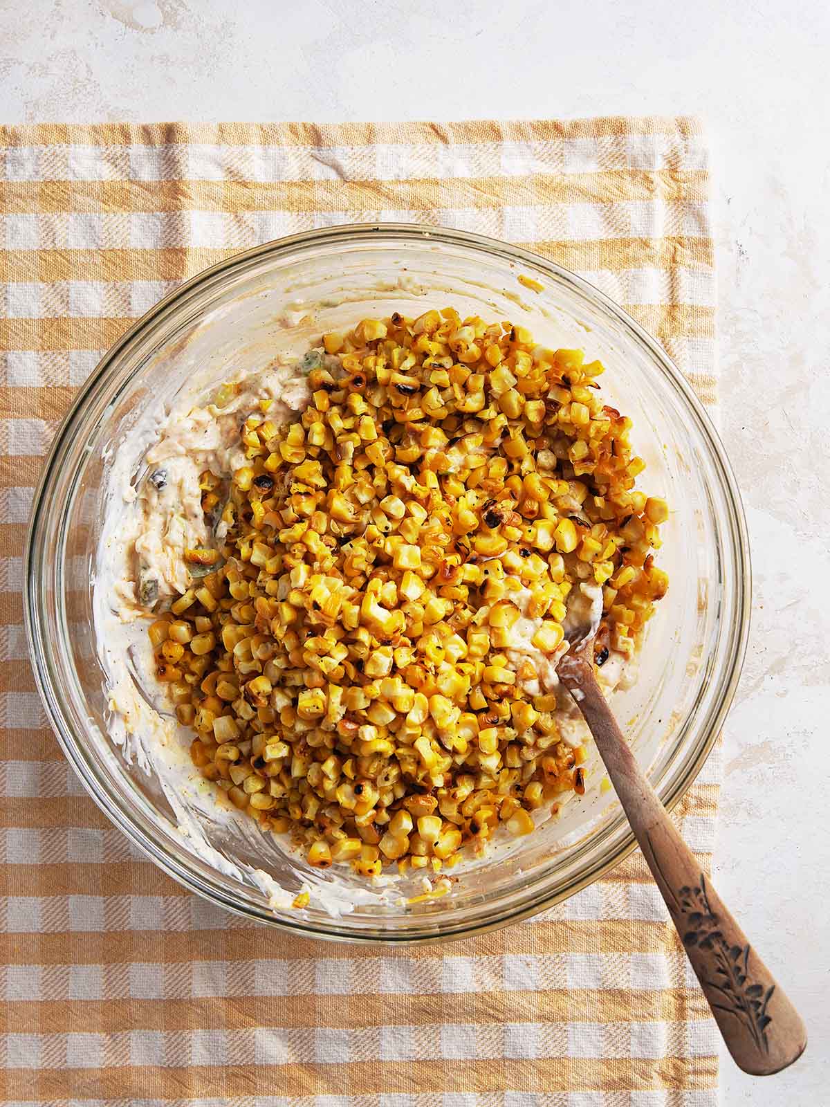 A glass bowl with cooked corn mixed into creamy ingredients sits on a beige checkered cloth. A wooden spoon rests in the bowl.