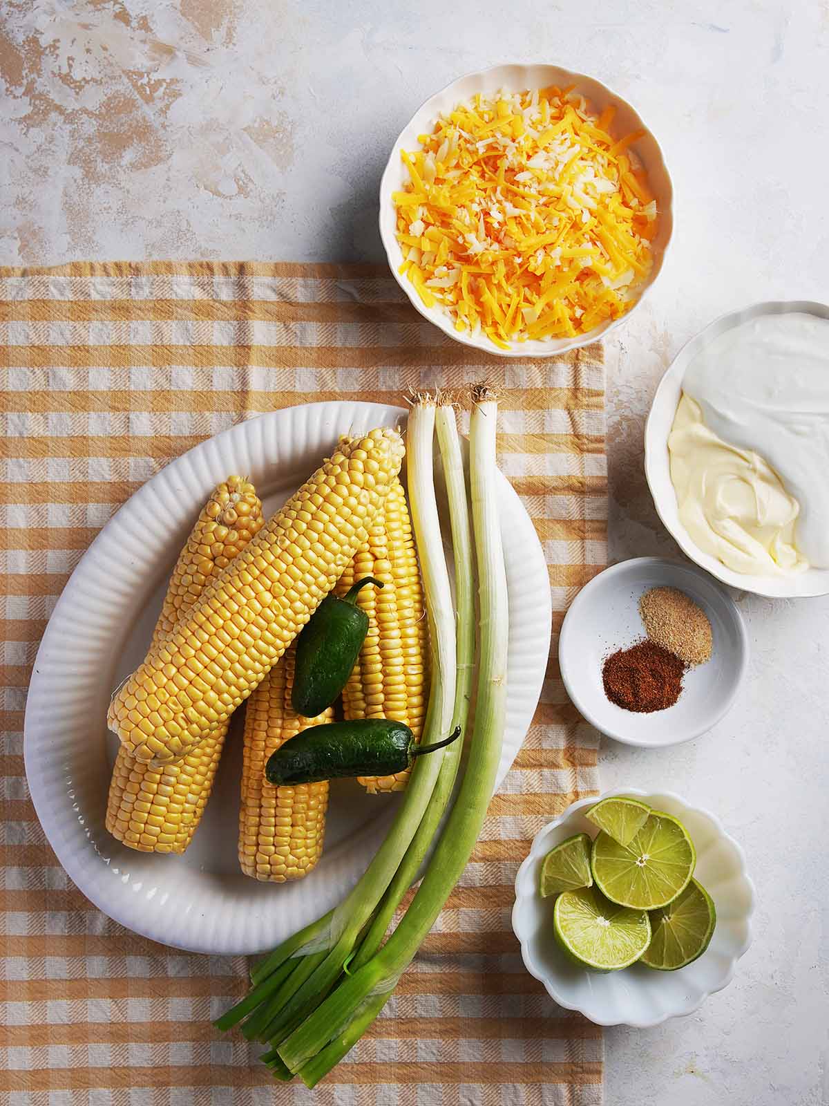 Overhead view of fresh corn, green onions, jalapeños, sliced limes, shredded cheese, spices, and a bowl of creamy sauce on a checkered cloth and white background.