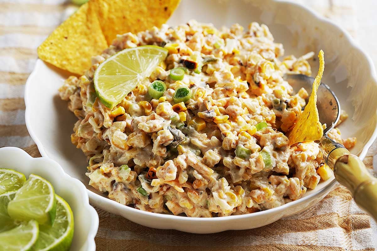 A Street Corn Dip with green onions, black beans, and peppers in a white bowl, garnished with lime wedges and tortilla chips. A spoon rests on the side and a small bowl of lime slices is nearby.