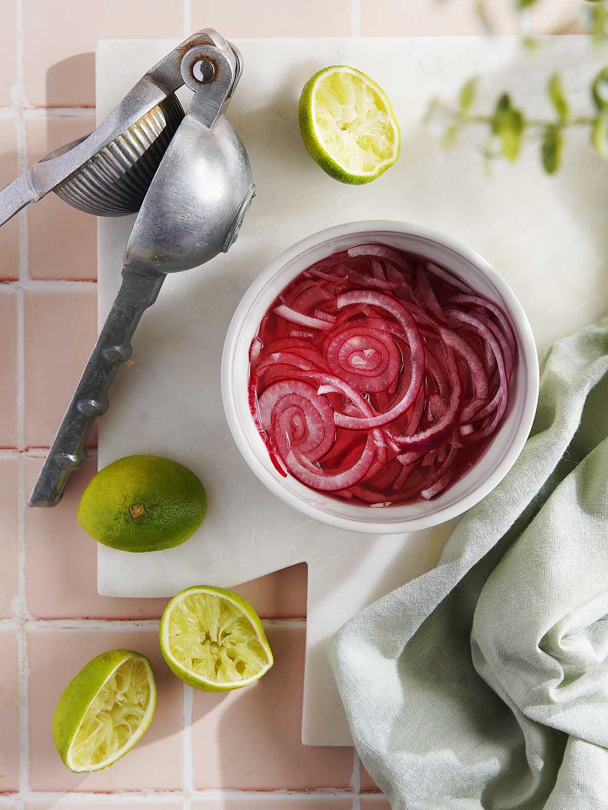 A small bowl of pickled red onions next to a metal citrus press and halved limes on a marble board.