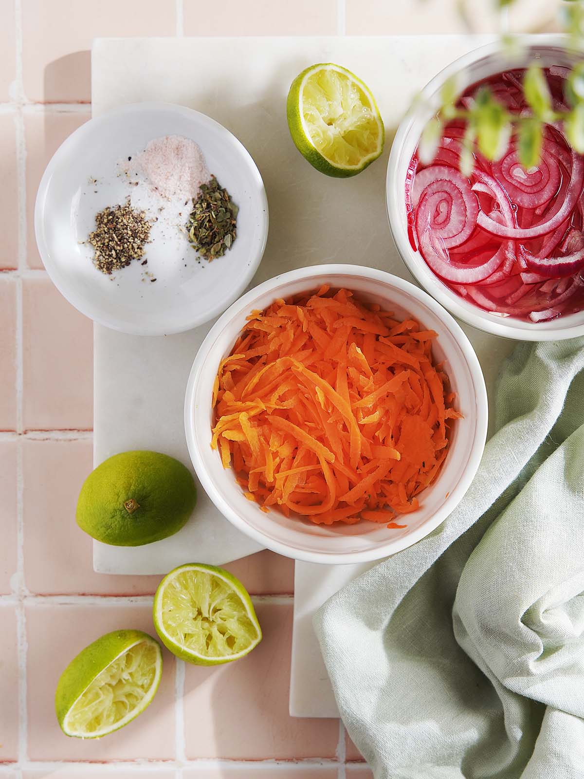 Bowls of shredded carrots, pickled red onions, and seasonings, surrounded by halved and whole limes.