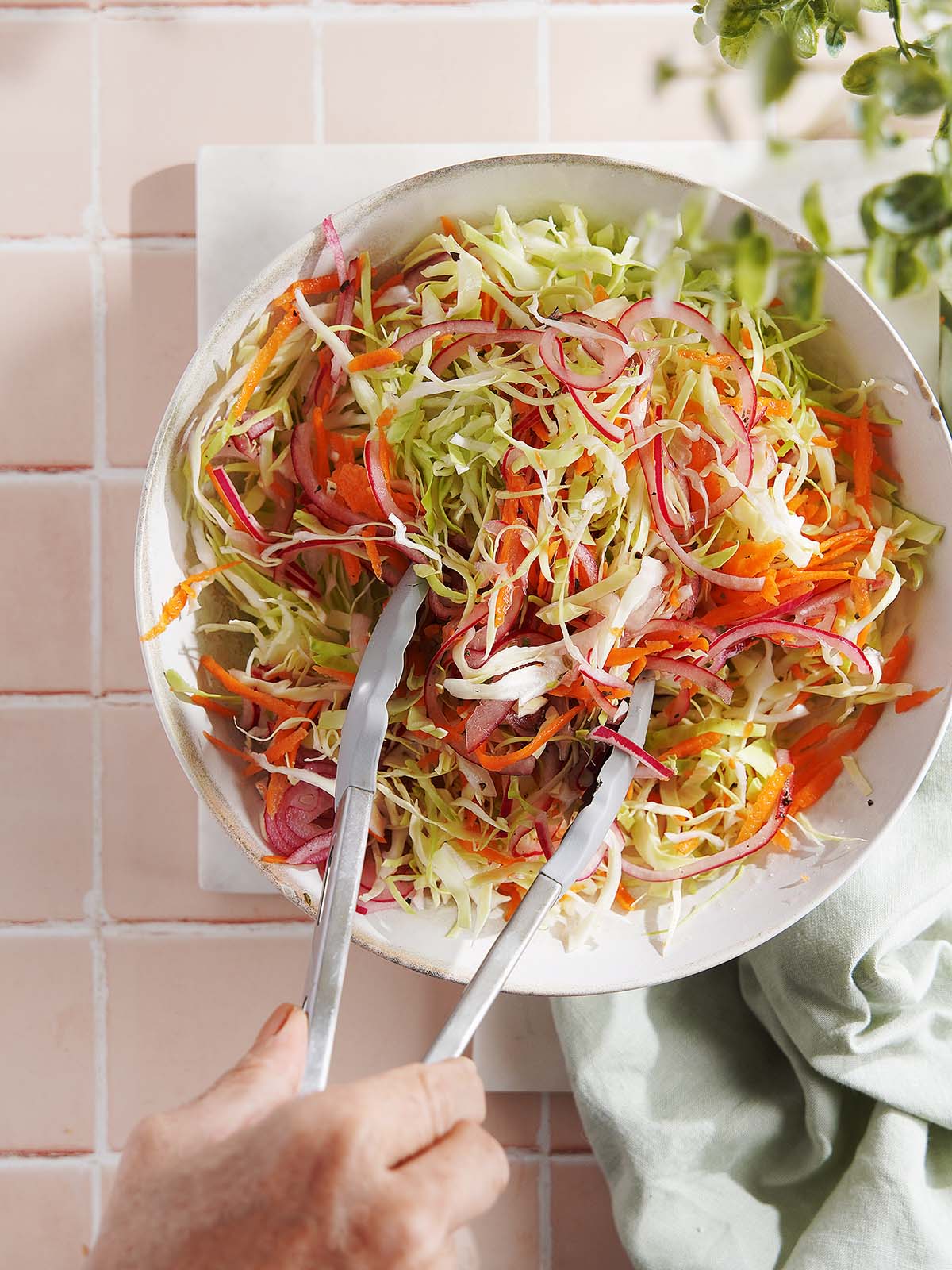 Hands using metal tongs to toss a bowl of coleslaw made with shredded cabbage, carrots, and red onion.