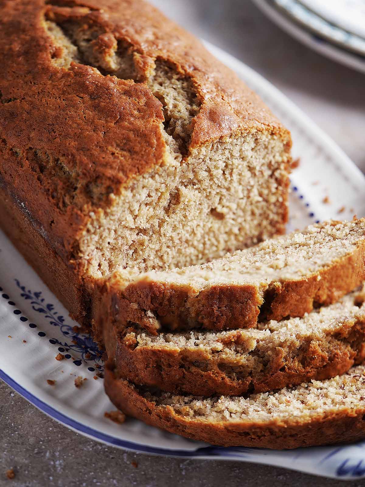 A loaf of banana bread on a patterned platter, with three slices cut and resting in front.