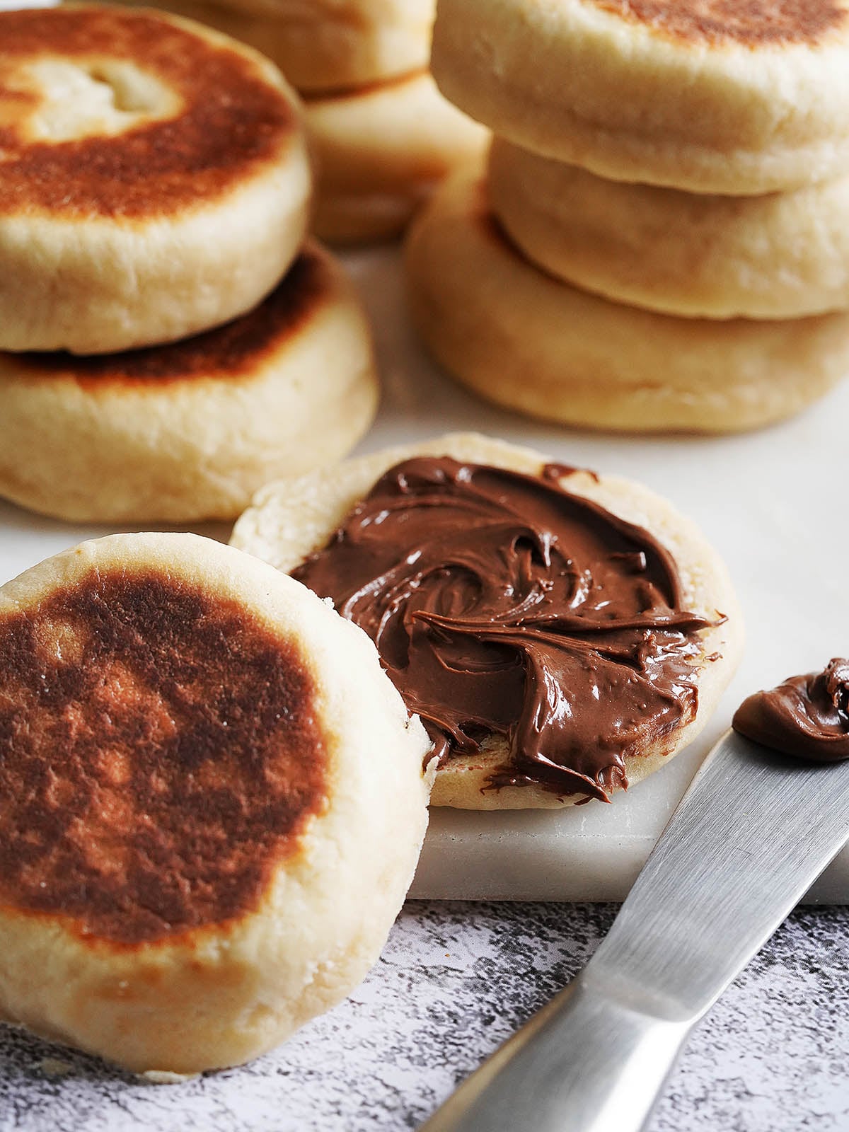 A halved Gordita De Nata with chocolate hazelnut spread, next to a butter knife and a stack of muffins.