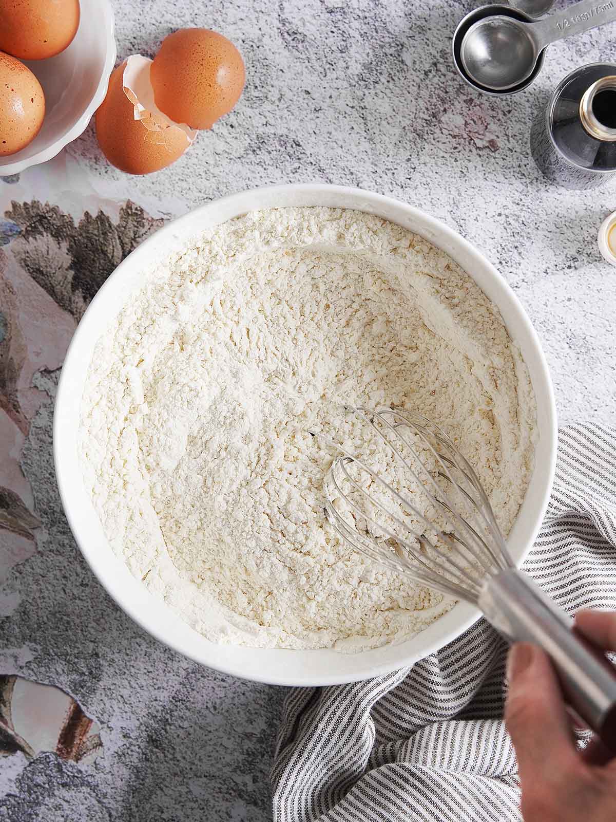 A hand uses a wire whisk to mix flour in a white bowl, with eggs and measuring spoons nearby.