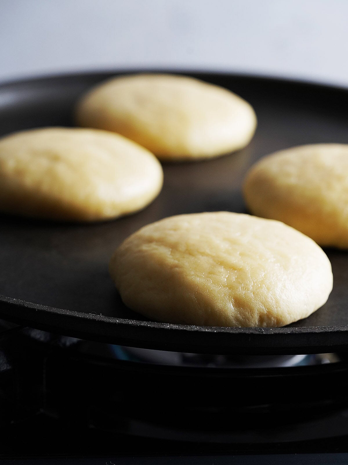 Four rounds of uncooked dough cooking on a black griddle over a blue gas flame.