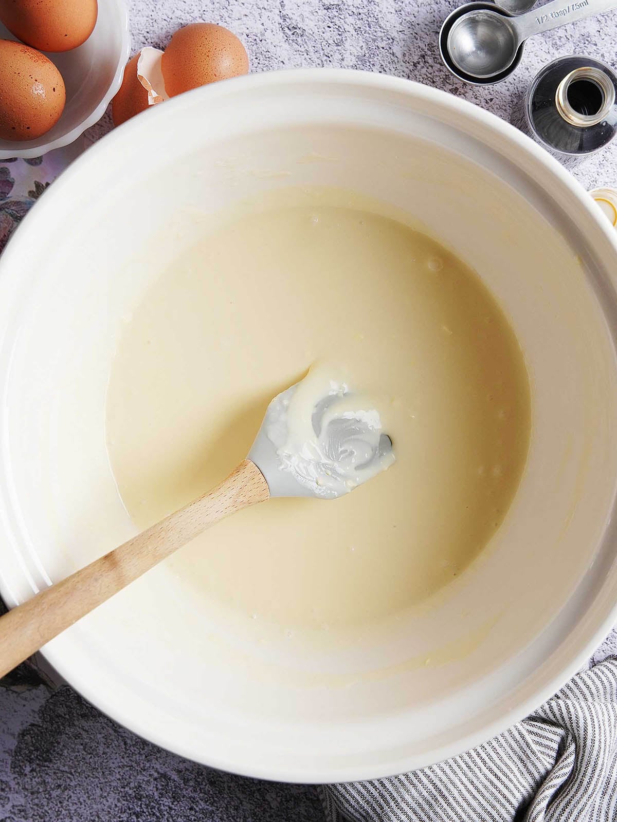 A silicone spatula rests inside a white mixing bowl filled with smooth cake or cookie batter, surrounded by eggs, measuring spoons, and vanilla extract.