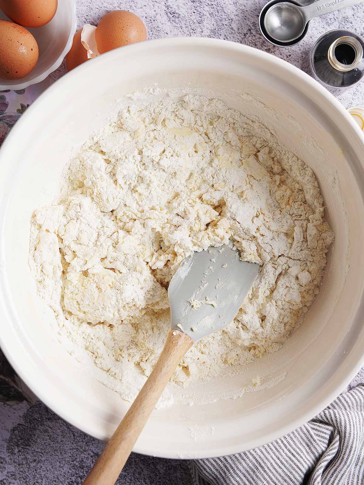 A silicone spatula mixing flour and ingredients in a white bowl, with eggs and measuring spoons nearby.