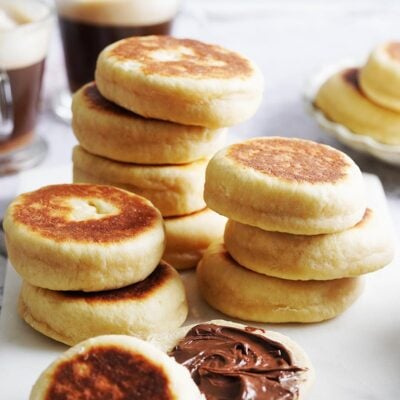 A stack of golden brown gorditas dulces on a marble board, with one muffin half spread with chocolate hazelnut spread.