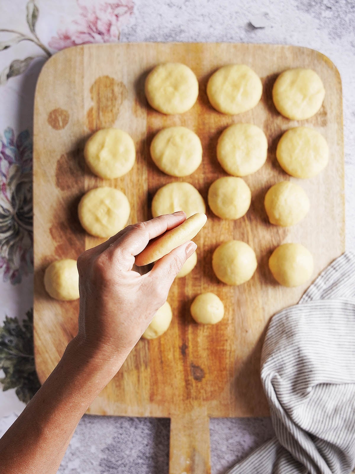 A hand holds a small round piece of dough over a wooden cutting board covered with rows of identical dough rounds.