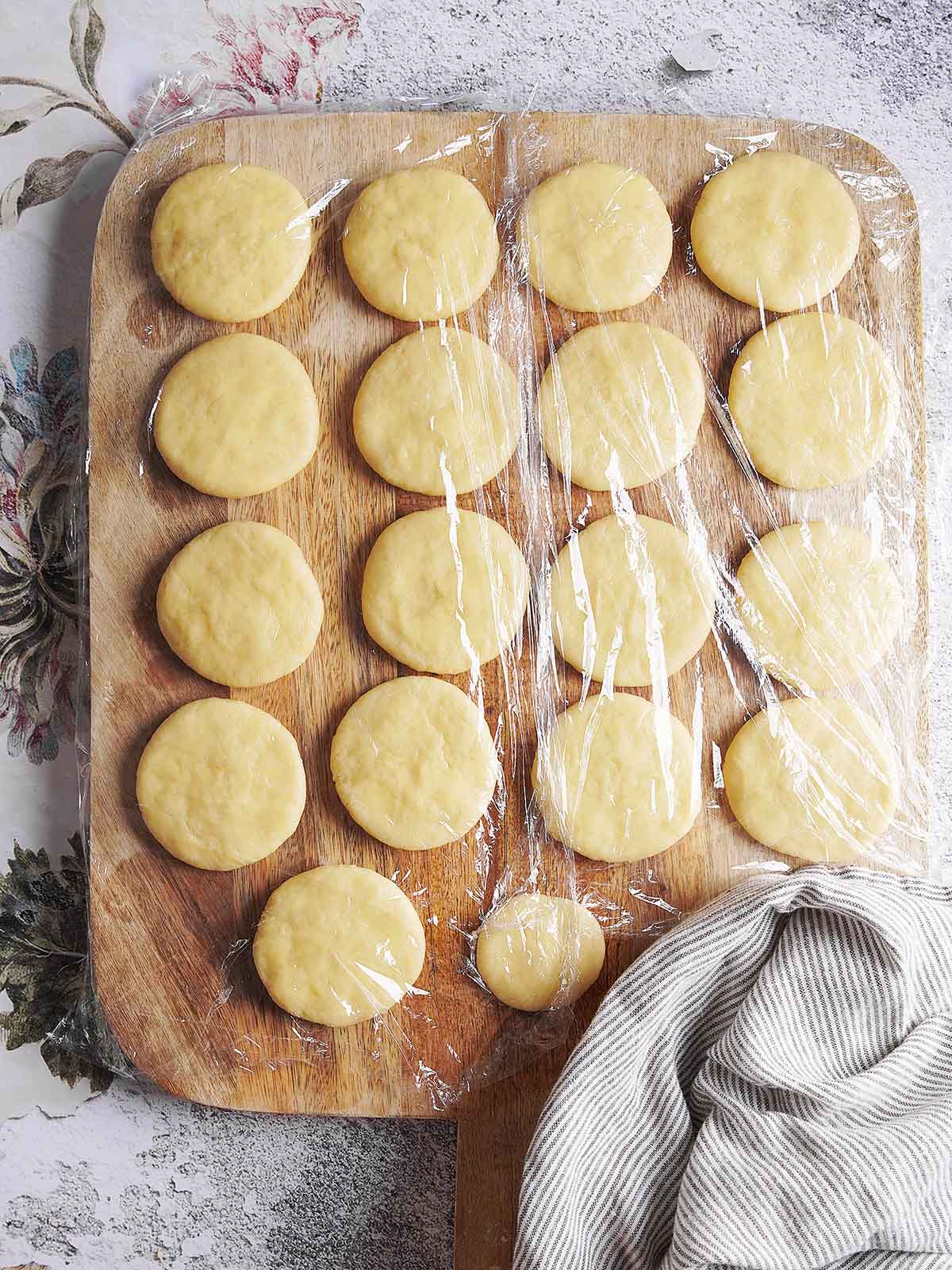 Seventeen rounds of raw dough resting on a wooden cutting board, covered with a sheet of plastic wrap.