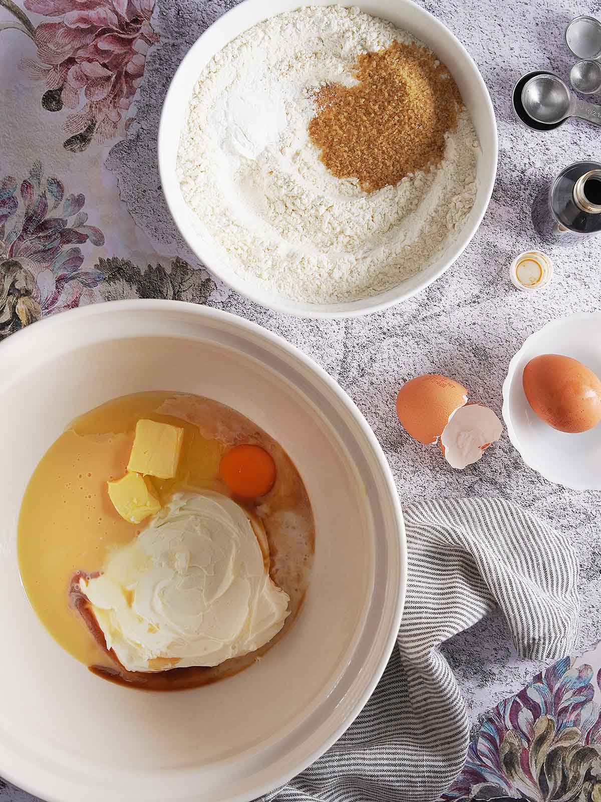 Two mixing bowls containing ingredients for baking, one with dry flour and brown sugar, and the other with butter, an egg, and cream or yogurt.