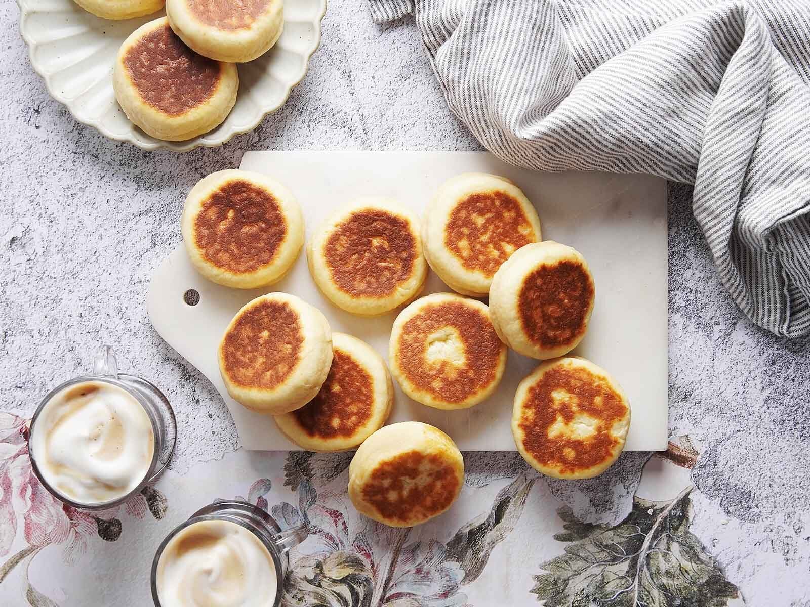 A marble board scattered with round, golden-brown griddle cakes, with a plate of more cakes, a folded napkin, and two cups of coffee nearby.
