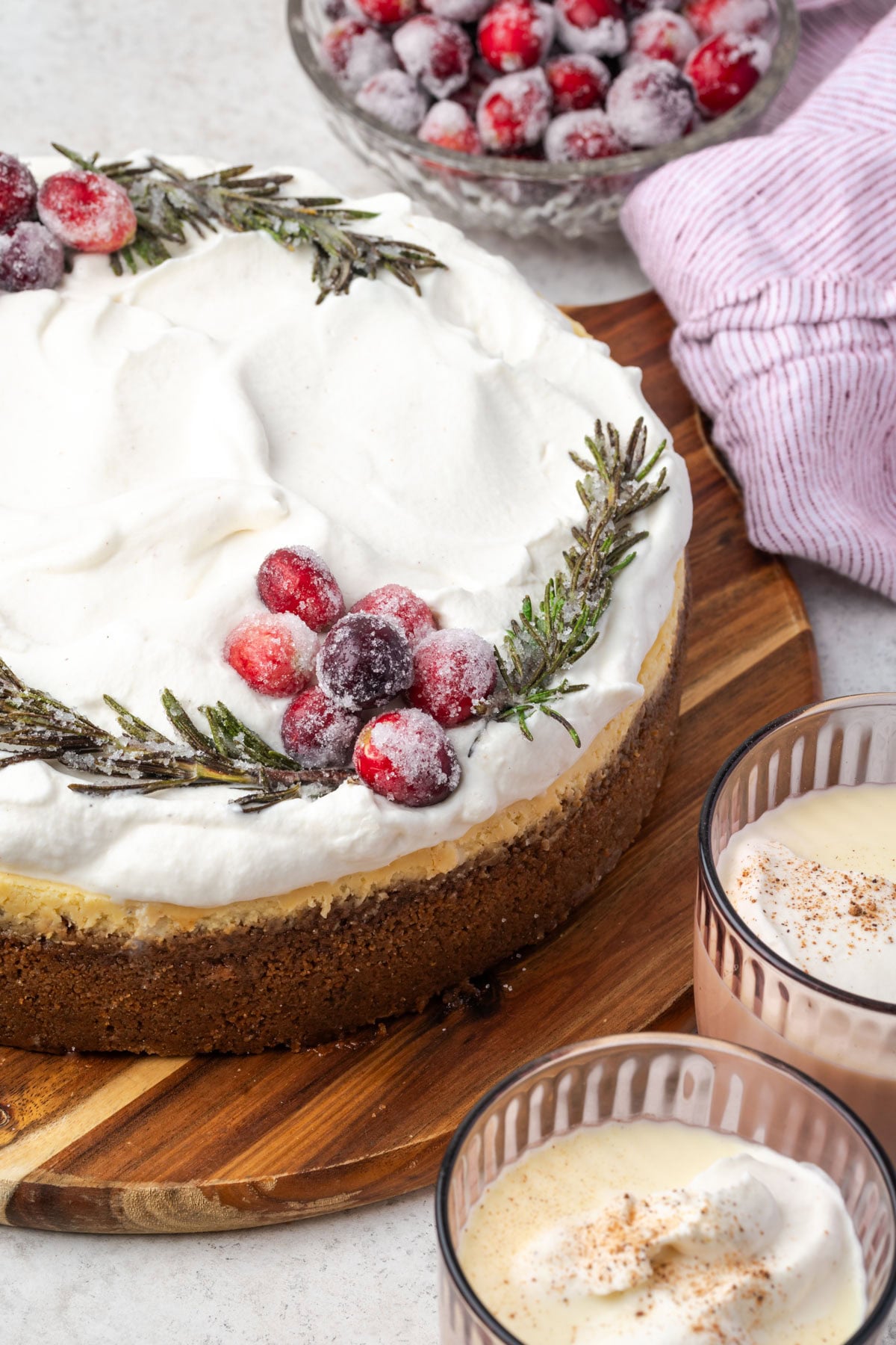 A cheesecake topped with whipped cream, sugared cranberries, and rosemary sprigs, with a bowl of cranberries and two glasses of eggnog nearby.