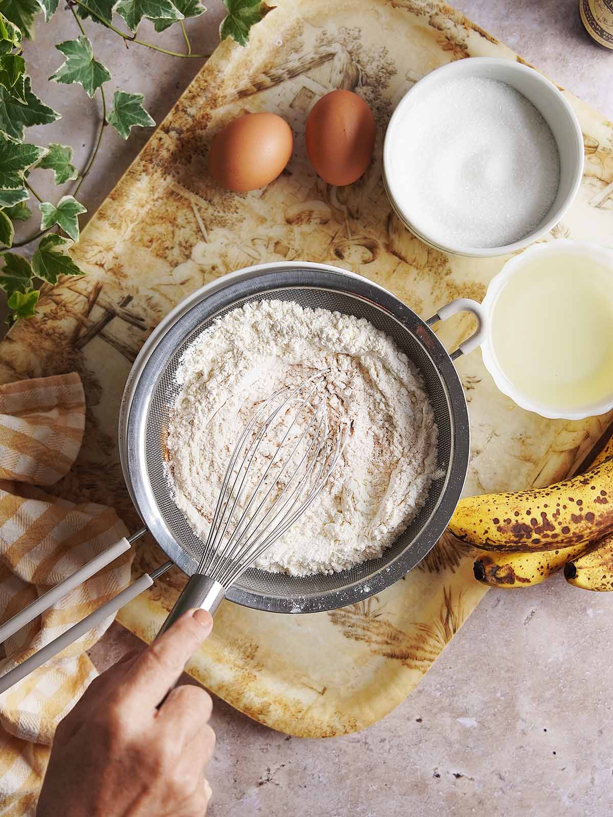 A hand whisks flour in a fine-mesh sieve over a patterned tray with two eggs, a bowl of sugar, a bowl of oil, and spotted bananas.