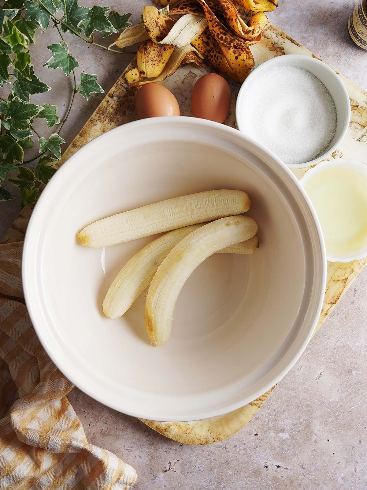 Three peeled bananas in a mixing bowl, surrounded by eggshells, two whole eggs, a small bowl of sugar, and a small bowl of oil.