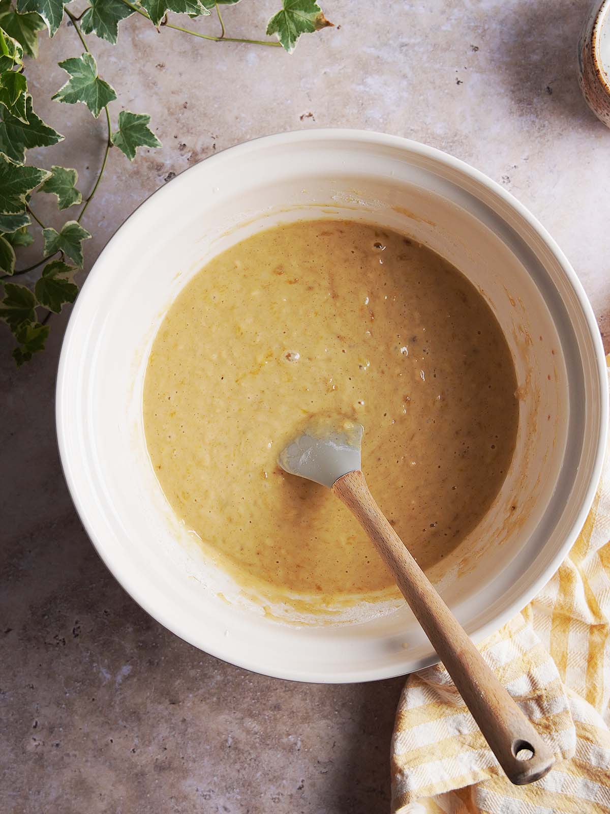 A silicone spatula mixing light brown cake or muffin batter in a large ceramic bowl.