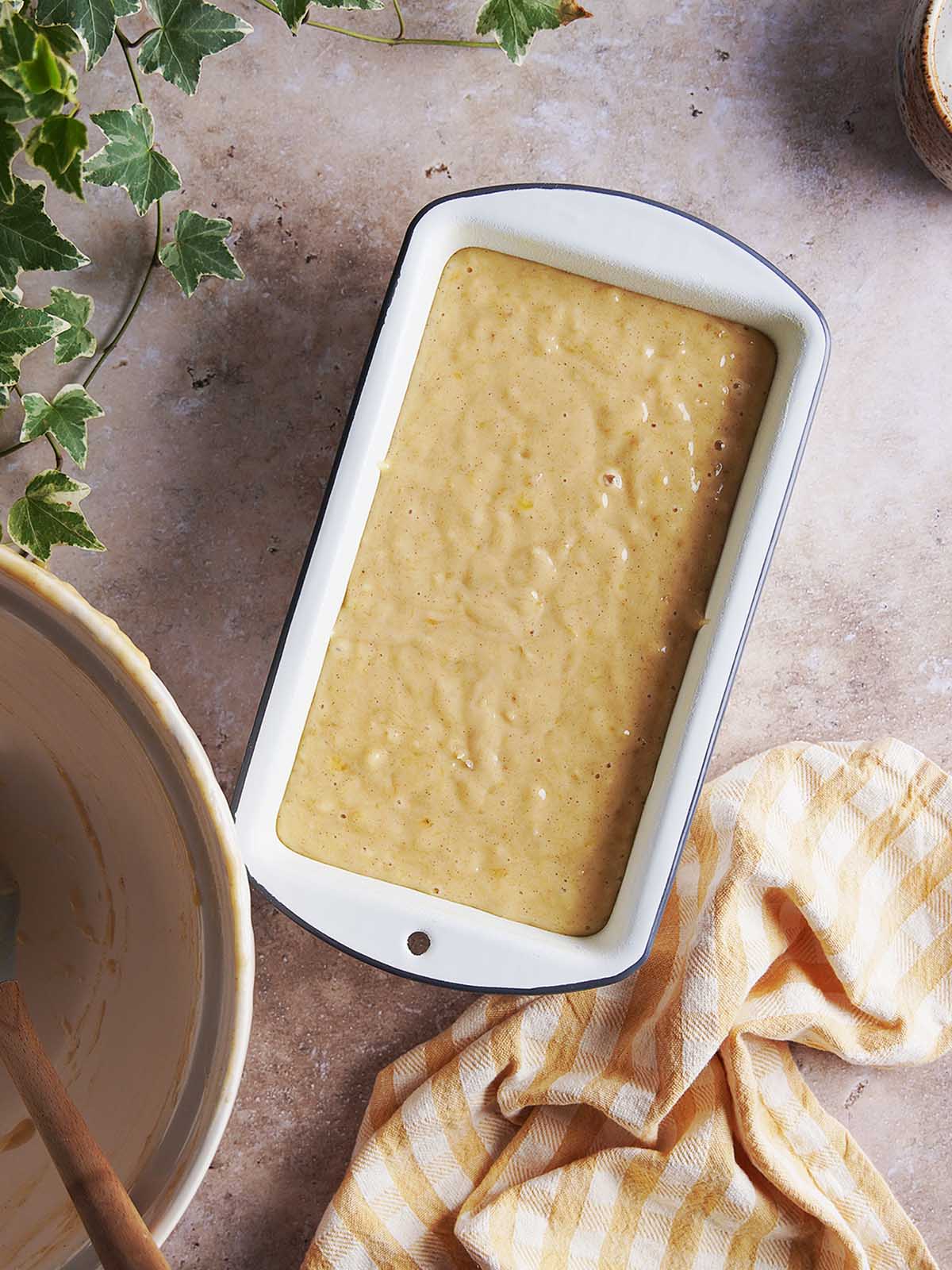 A loaf pan filled with smooth batter next to a mixing bowl and a yellow checkered towel.